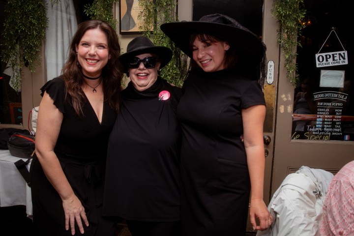 Three women in black dresses and hats, smiling indoors.