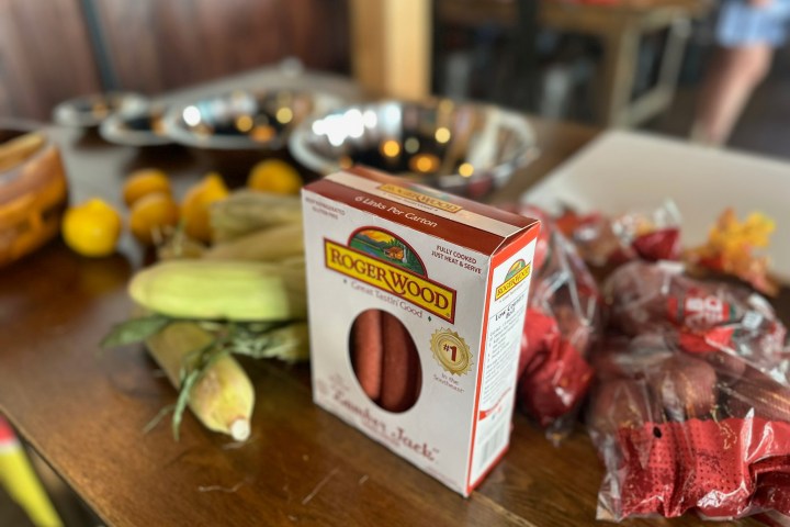 Packaged sausages, corn, and citrus on a wooden table, with blurred background.