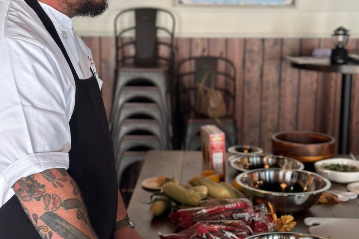 Chef with tattoos slices lemon on a wooden table with various ingredients.