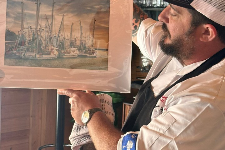 Man in a cap and apron holding a framed photo of boats in a harbor.
